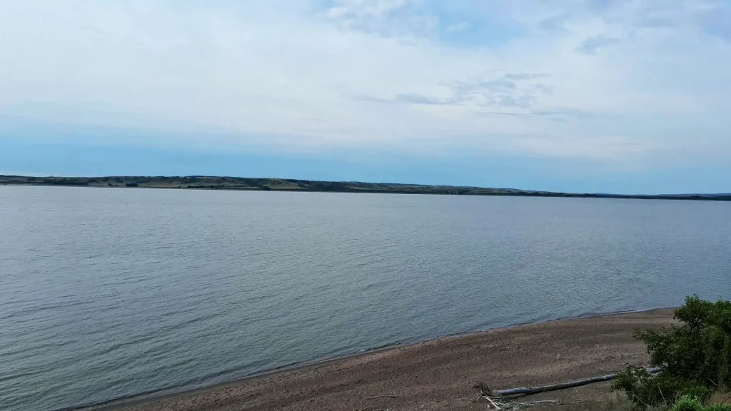A serene lake scene featuring calm water, soft sand, and lush green trees lining the shore.