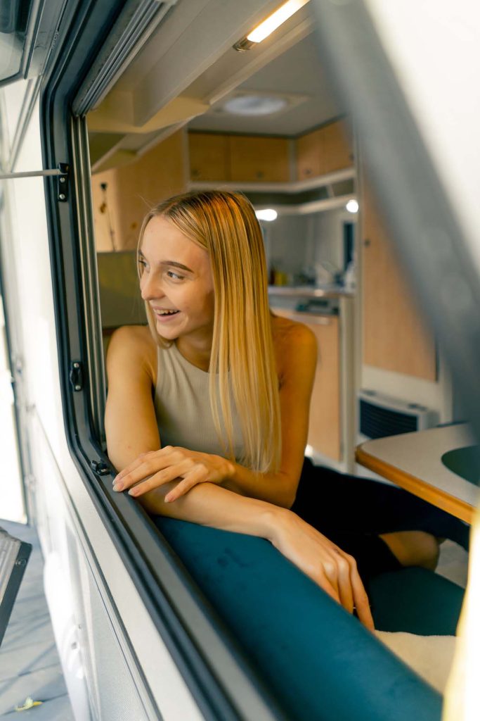 A woman sits in the window of a camper van, enjoying the view outside with a relaxed expression.